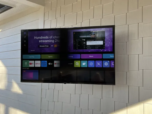A TV mounted on a white shake-shingle wall under a vaulted wood porch ceiling