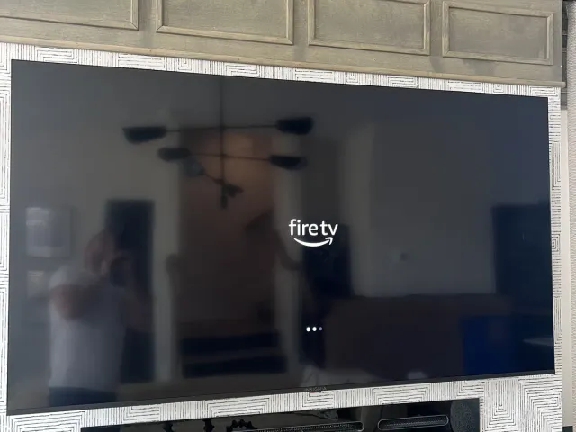 A TV mounted on a textured accent wall above a linear electric fireplace, under gray coffered paneling