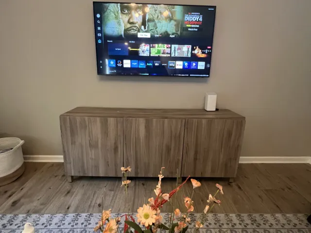 A TV on a flat mount on a beige wall above a wood console with flowers beside it