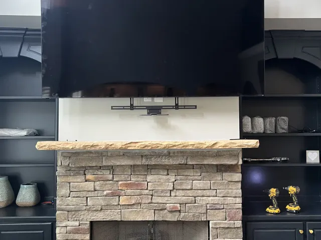 A TV mounted above a live-edge wood mantel on a stacked-stone fireplace with black built-in shelving