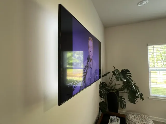 A TV on a tilt mount on a white bedroom wall beside a window