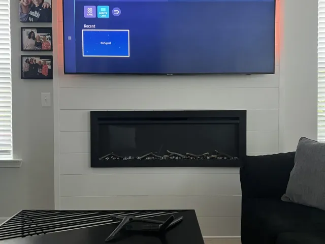 A TV mounted on a white shiplap wall above a linear electric fireplace, a black sectional in front