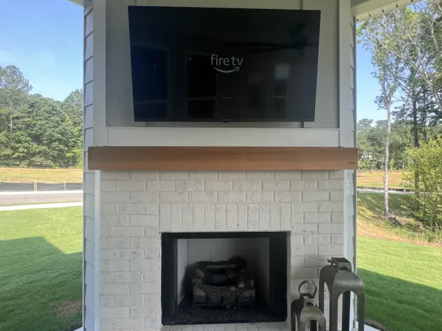 A TV mounted above a wood mantel on a white painted-brick fireplace on a covered porch, lawn and trees behind