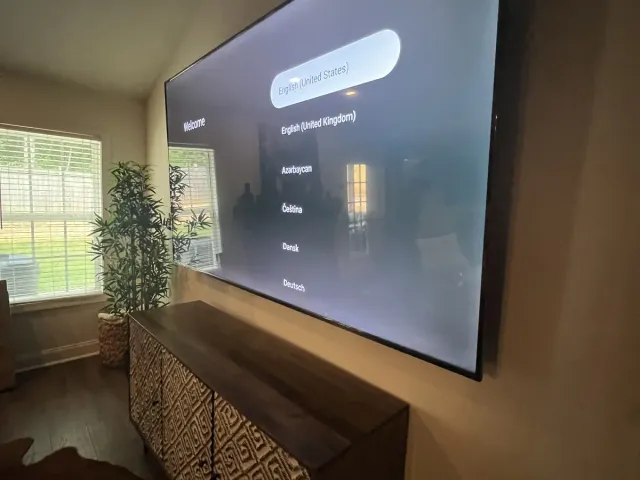 A TV on a gray bedroom wall, a plant in the corner by the window