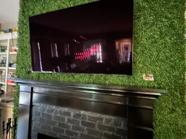 A TV mounted on an artificial-greenery accent wall above a black mantel and black brick fireplace