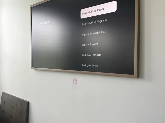 A thin-bezel TV mounted flush on a white wall, the language setup screen showing