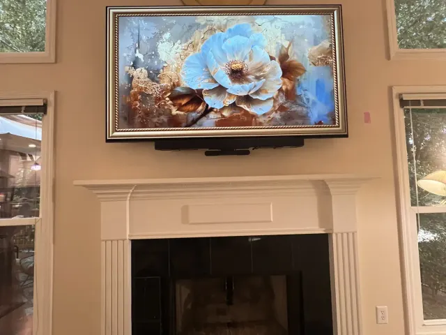 A Samsung Frame TV with an ornate bezel showing a floral artwork, a soundbar beneath it, above a white mantel with a black tile surround