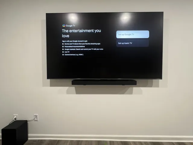 A TV with a soundbar under it on a white wall near a wood-floor hallway