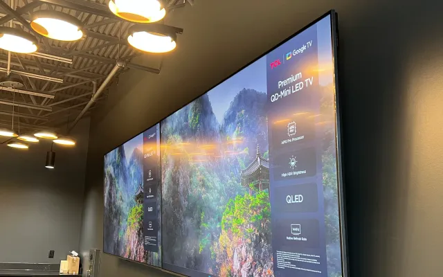Two large TVs mounted side by side on a dark wall under industrial ceiling lights